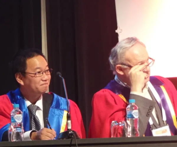 Dr Peter Kim in academic dress seated at a conference panel table beside a colleague